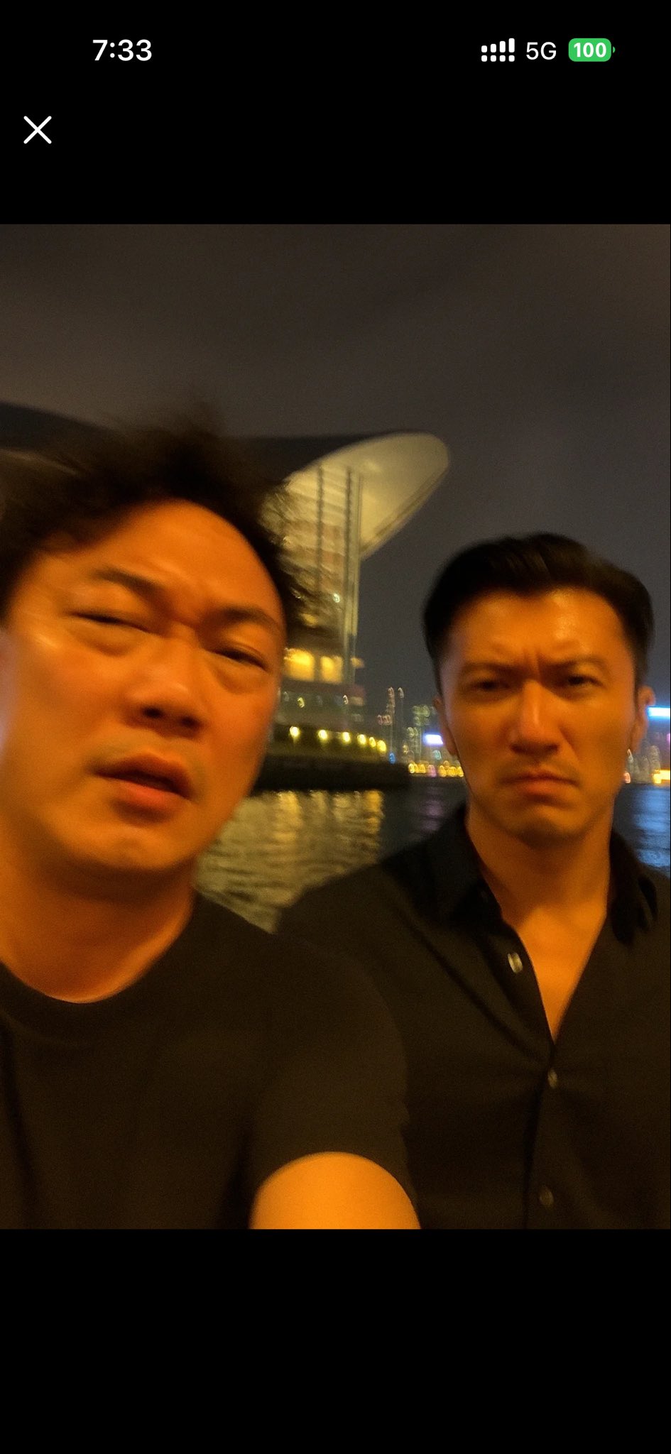 请画一张极其平凡无奇的iPhone 自拍照,没有明确的主体或构图感,就像是随手一拍的快照。照片略带运动模糊,阳光或店内灯光不均导致轻微曝光过度。角度尴尬、构图混乱,整体呈现出一种刻意的平庸感-就像是从口袋里拿手机时不小心拍到的一张自拍。主角是陈奕迅和谢霆锋,晚上,旁边是香港会展中心,在香港维多利亚港旁边。